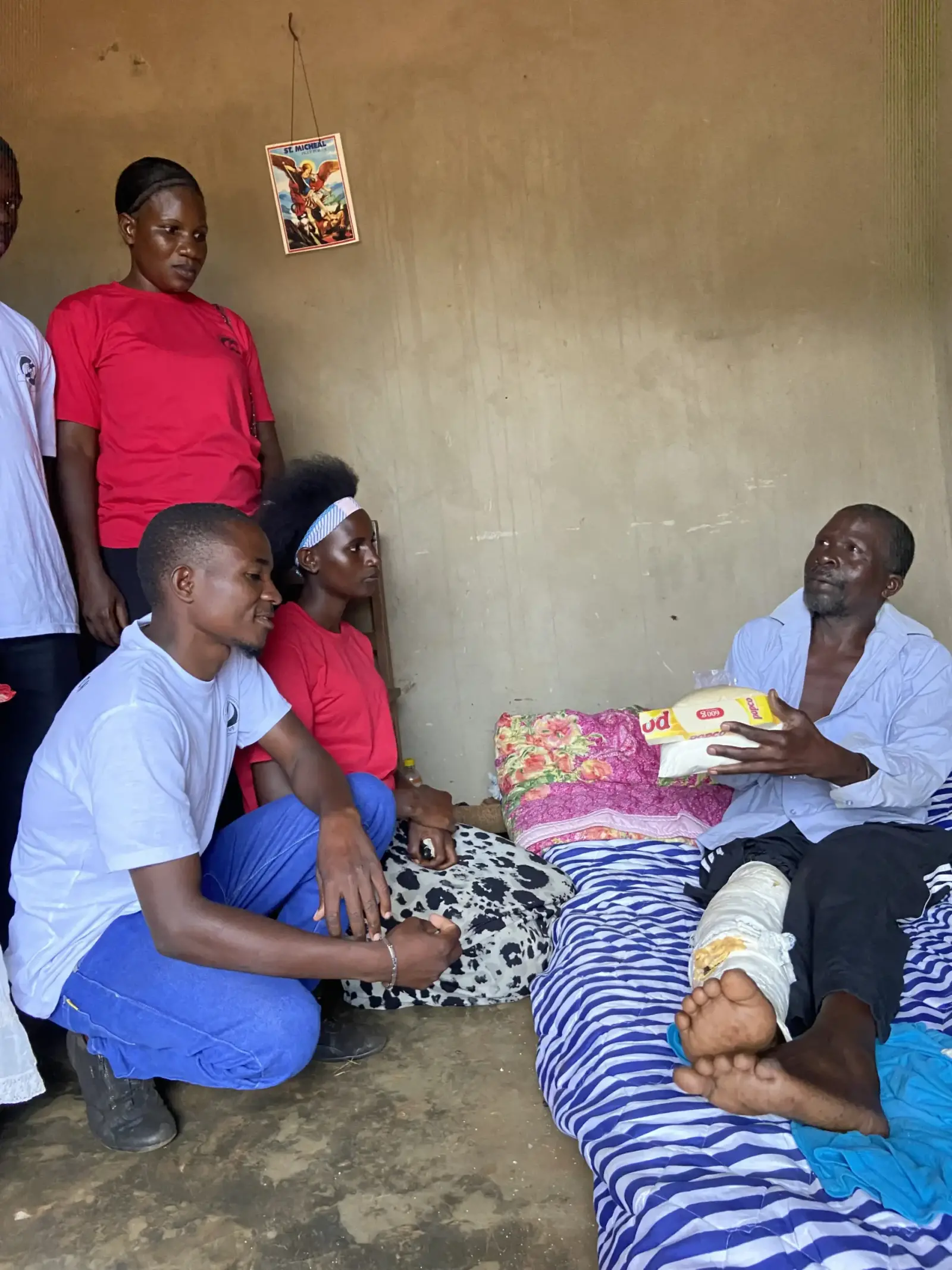 Volunteers in red shirts visiting an elderly man resting on a mattress at home